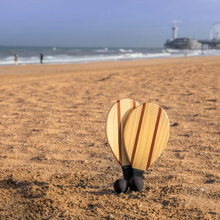 Outdoor wooden Frescobol tennis set by XD Collection with two wooden paddles and a ball on a sandy outdoor surface.